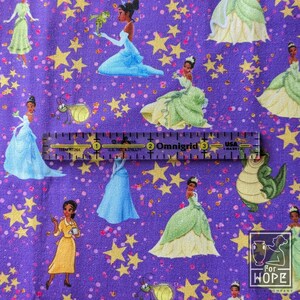 May include: Purple fabric with a repeating pattern of a princess in various gowns, frogs, and stars. A ruler is placed across the fabric. The text "Omnigrid" and "For Hope Company" are visible.