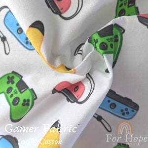May include: White fabric with a repeating pattern of colorful video game controllers and headsets. The fabric has the text "Gamer Fabric 100% Cotton" and "For Hope Waco Texas" printed on it.