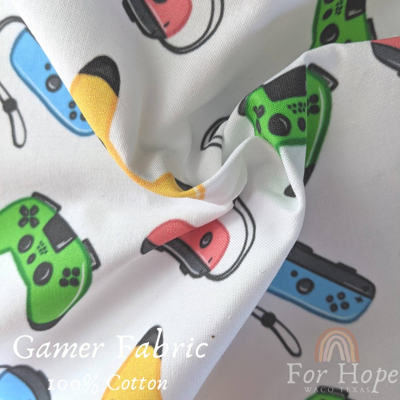 May include: White fabric with a repeating pattern of colorful video game controllers and headsets. The fabric has the text "Gamer Fabric 100% Cotton" and "For Hope Waco Texas" printed on it.