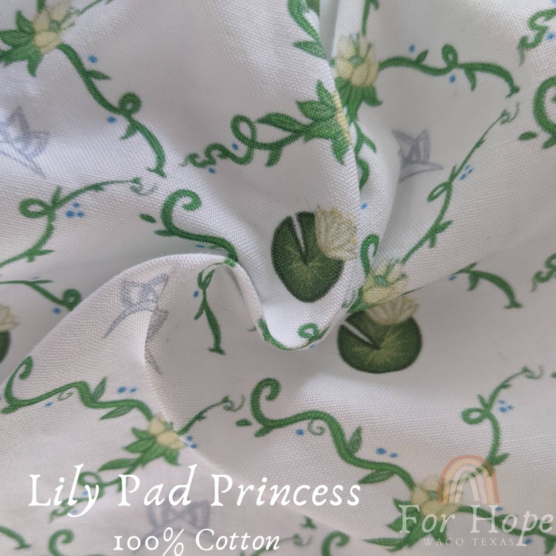 Peut inclure: Tissu en coton blanc avec un motif r&eacute;p&eacute;t&eacute; de n&eacute;nuphars verts, de fleurs blanches et de vignes. Le tissu porte l'inscription "Lily Pad Princess" et "For Hope".