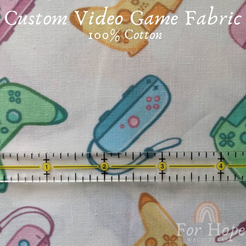 May include: A white cotton fabric with a repeating pattern of colorful video game controllers. The fabric is 100% cotton and has a ruler measuring 4 inches across the bottom of the image.