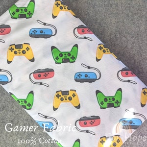 Könnte beinhalten: Weißer Baumwollstoff mit einem sich wiederholenden Muster aus bunten Videospielcontrollern. Der Stoff trägt den Text "Gamer Fabric 100% Cotton".