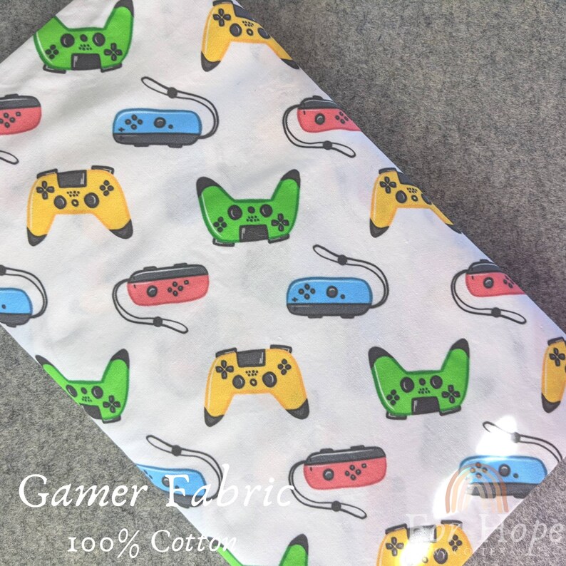 May include: White cotton fabric with a repeating pattern of colorful video game controllers. The fabric has the text "Gamer Fabric 100% Cotton" printed on it.