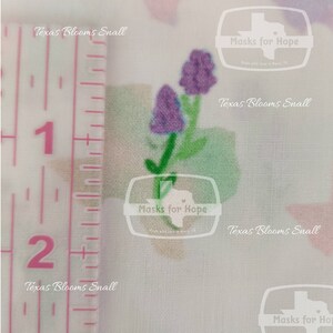 May include: A close-up of a white fabric with a purple flower design. The fabric is measured with a ruler showing 1 and 2 inches. The fabric has the text "Texas Blooms Small" and "Masks for Hope" printed on it.