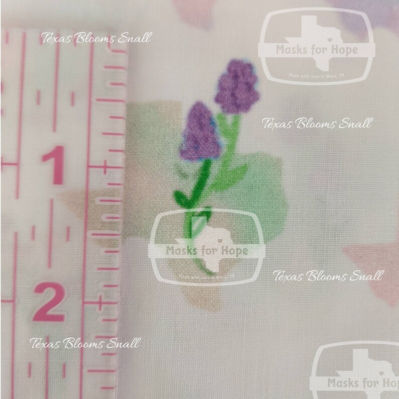 May include: A close-up of a white fabric with a purple flower design. The fabric is measured with a ruler showing 1 and 2 inches. The fabric has the text "Texas Blooms Small" and "Masks for Hope" printed on it.