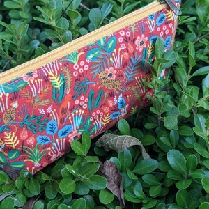 K&ouml;nnte beinhalten: Eine rote Rei&szlig;verschlusstasche mit einem gelben Rei&szlig;verschluss und einem bunten Dschungelprint mit Blumen, Bl&auml;ttern und Tieren.