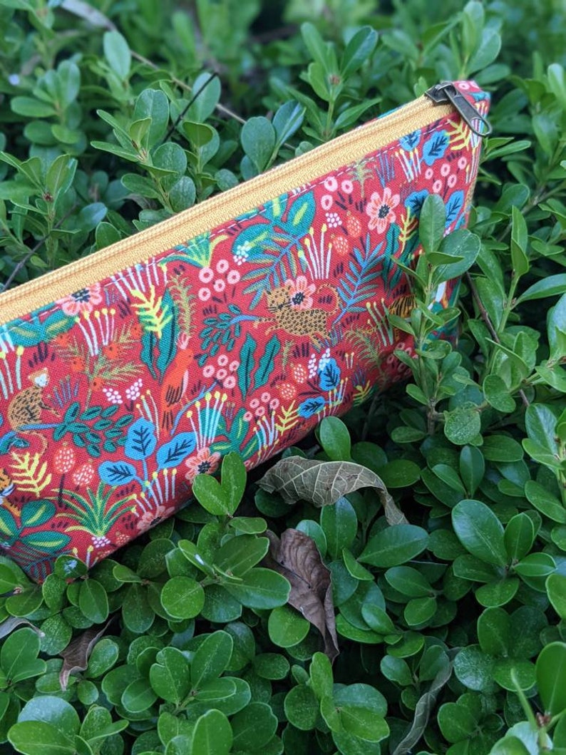 K&ouml;nnte beinhalten: Eine rote Rei&szlig;verschlusstasche mit einem gelben Rei&szlig;verschluss und einem bunten Dschungelprint mit Blumen, Bl&auml;ttern und Tieren.