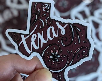 May include: A 3 inch die cut sticker featuring a burgundy outline of the state of Texas with a paisley pattern and the word "Texas" written in white script.