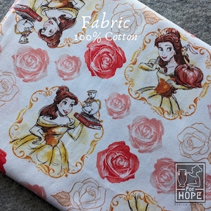 May include: White fabric with a repeating pattern featuring Belle from Beauty and the Beast, red roses, and gold frames. The fabric also has the text "Fabric 100% Cotton". The design is suitable for various sewing and crafting projects.