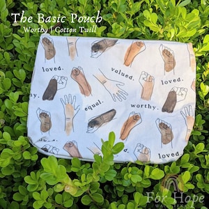 May include: A white cotton pouch with a repeating pattern of brown and black hands in a fist shape. The words "loved", "valued", "equal", and "worthy" are printed on the pouch. The pouch is labeled "The Basic Pouch" and "Worthy Cotton Twill".