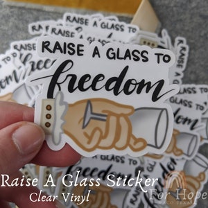 May include: A white sticker with a hand raising a glass and the text "Raise a glass to freedom". The sticker is made of clear vinyl.