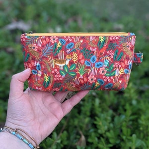 K&ouml;nnte beinhalten: Eine rote Rei&szlig;verschlusstasche mit einem bunten Dschungel-Druck mit Tigern, V&ouml;geln und Blumen. Die Tasche hat einen gelben Rei&szlig;verschluss und einen gelben Zipper.