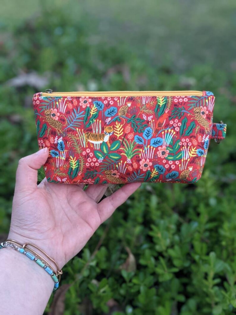 K&ouml;nnte beinhalten: Eine rote Rei&szlig;verschlusstasche mit einem bunten Dschungel-Druck mit Tigern, V&ouml;geln und Blumen. Die Tasche hat einen gelben Rei&szlig;verschluss und einen gelben Zipper.