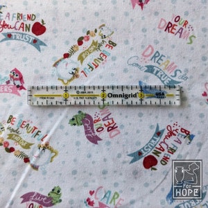 May include: White fabric with a repeating pattern of whimsical illustrations, banners, and text. The text includes phrases like "Be Beautiful" and "A Friend You Can Trust." A clear ruler with inch markings is laid across the fabric.