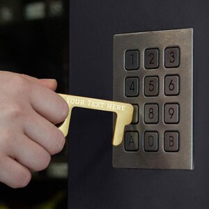 May include: A gold-colored, multi-purpose tool with the text "YOUR TEXT HERE" engraved on it. The tool is being used to press a button on a keypad with the numbers 1 through 9 and the letters A, O, and B.