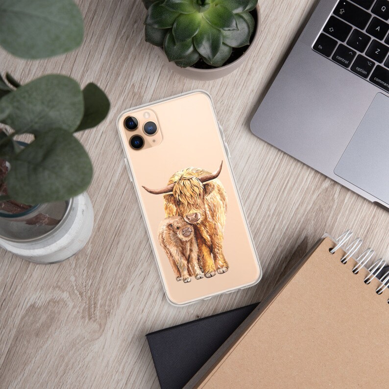 May include: A clear phone case with a watercolor illustration of a Highland cow and her calf. The cow is brown and white with long, shaggy hair. The calf is smaller and has a more reddish-brown color.