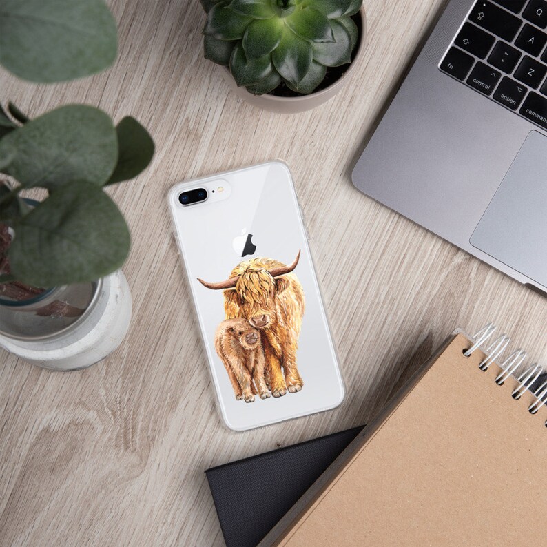 May include: A clear phone case with a watercolor illustration of a Highland cow and her calf. The cow is brown and white with long, shaggy hair. The calf is smaller and has a more reddish-brown color.