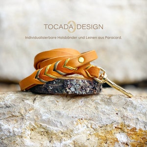 May include: A tan leather dog leash with a gold-colored clasp and a decorative braided section with green, yellow, and orange accents. The leash is coiled on a piece of wood, with the text "TOCADA DESIGN" and "Individualisierbare Halsbänder und Leinen aus Paracord."