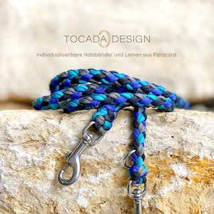 May include: A braided paracord dog leash with two silver clips. The leash is blue, teal, black, and gray. The text "TOCADADESIGN" and "Individualisierbare Halsbänder und Leinen aus Paracord." is visible in the image.