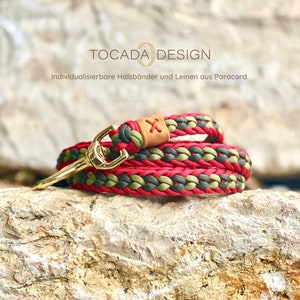 May include: Red, green, and brown paracord dog leash with a gold metal clasp. The leash is braided and has a leather patch with an X on it. The text "TOCADADESIGN" and "Individualisierbare Halsbänder und Leinen aus Paracord." is visible in the background.