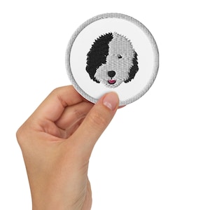 Puede incluir: Un parche bordado con la cara de un perro, con pelo blanco y negro y una nariz rosa.