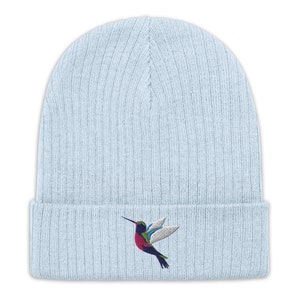 Op de afbeelding: Een lichtblauwe gebreide beanie met een kleurrijke geborduurde kolibrie op de voorkant.