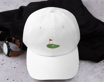 Op de afbeelding: Witte baseballpet met een geborduurd groen golfveld met een rode vlag en een witte golfbal.