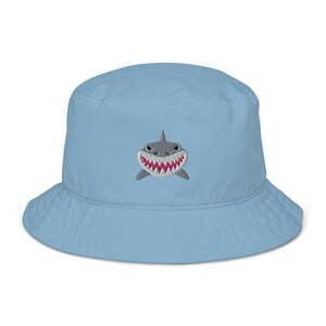 May include: A light blue bucket hat with an embroidered shark face on the front. The shark has a white body, gray eyes, and a pink open mouth with sharp teeth.