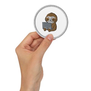May include: A round white embroidered patch with a gray border featuring a brown sloth wearing glasses and using a laptop.