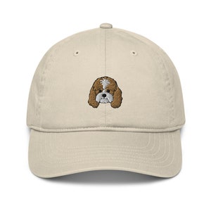 Puede incluir: Una gorra de béisbol beige con un rostro de perro bordado en marrón y blanco.