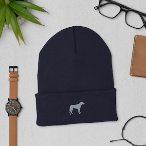 May include: A navy blue beanie with a light gray embroidered dog silhouette. The hat is made of a soft, knitted material. The beanie is displayed on a light gray surface with a watch, glasses, and a wallet.