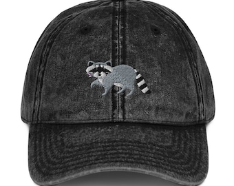 May include: A black vintage washed denim baseball cap with an embroidered raccoon design.