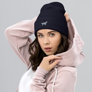 May include: A navy blue beanie with a gray embroidered silhouette of a dog. The beanie is being worn by a person with long brown hair.