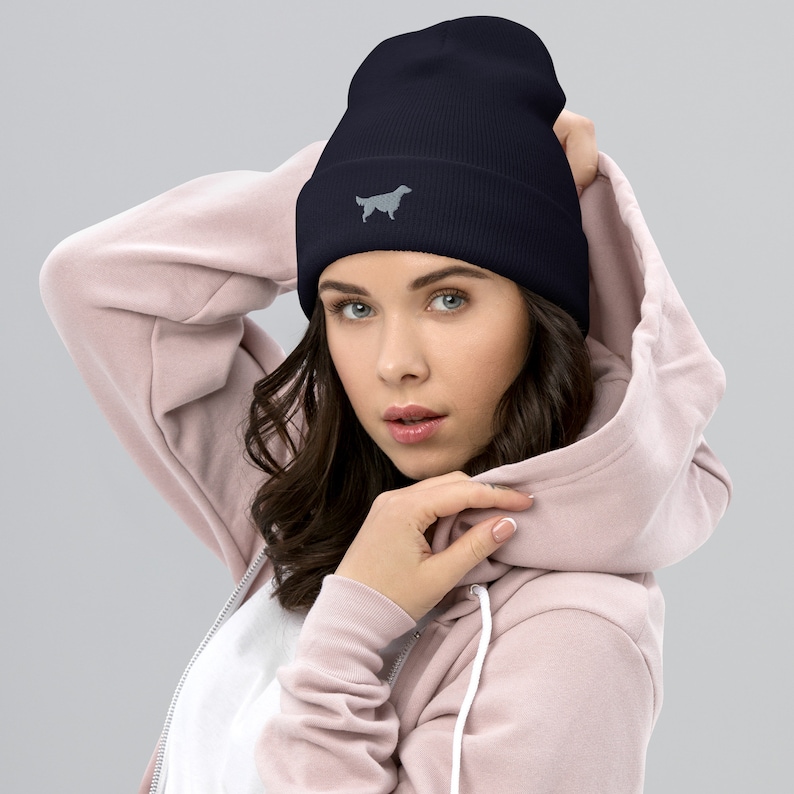 May include: A navy blue beanie with a gray embroidered silhouette of a dog. The beanie is being worn by a person with long brown hair.