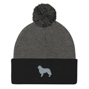 May include: A gray and black knit beanie with a fluffy pom pom on top. The beanie has a silhouette of a dog embroidered on the front.