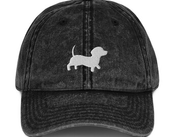 Puede incluir: Gorra de béisbol negra desgastada con un diseño de perro salchicha bordado en blanco. La gorra tiene una visera curva y una correa ajustable. El sombrero está hecho de un material suave y desgastado, lo que le da un aspecto vintage.