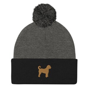 May include: A gray and black knit beanie with a fluffy pom pom on top. The beanie has a brown embroidered silhouette of a dog on the front.