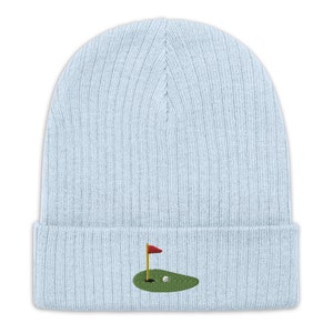 Op de afbeelding: Een lichtblauwe gebreide beanie met een klein geborduurd golfveld met een vlag en een golfbal.