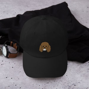 Puede incluir: Una gorra de béisbol negra con un rostro de caniche marrón bordado en la parte delantera.