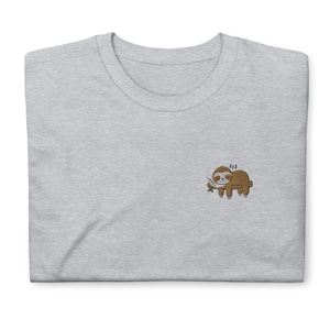 Könnte beinhalten: Ein graues T-Shirt mit einem gestickten Bild eines schlafenden braunen Faultiers auf einem Ast mit einem Kissen. Das Faultier hat ein kleines Lächeln und geschlossene Augen. Der Text "zzz" befindet sich über dem Faultier.