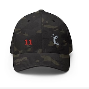 Peut inclure: Une casquette de baseball camouflage noire et verte avec un graphisme de joueur de volley-ball blanc et le numéro "11" en rouge.