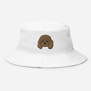Puede incluir: Sombrero de pescador blanco con un rostro de perro marrón bordado. El sombrero tiene un diseño casual, ideal para la protección solar. La cara del perro está centrada en la parte delantera, añadiendo un toque lúdico al accesorio.