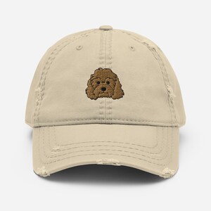 May include: A beige distressed baseball cap with an embroidered brown poodle on the front.