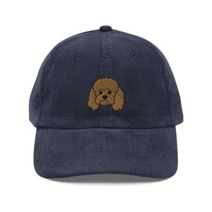 Puede incluir: Una gorra de béisbol de pana azul marino con un rostro de caniche marrón bordado.