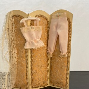 May include: A miniature three-panel screen with a gold and white floral pattern. A pink lace-trimmed camisole and matching pants hang on the screen.