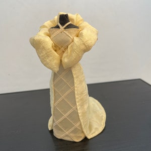 May include: A small, cream-colored dress form with a white dress with a diamond pattern. The dress has a large, ruffled collar.