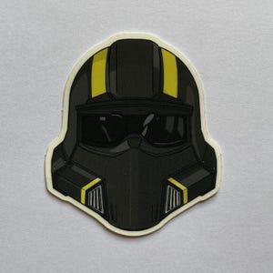 以下が含まれることがあります： バイザー付きの黒と黄色のヘルメットステッカー。ヘルメットは中央に黄色のストライプがあり、側面に黄色のアクセントがあります。