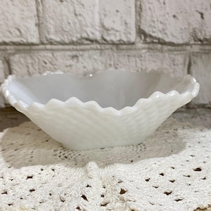 May include: White milk glass bowl with a scalloped edge, sitting on a white crocheted doily on a wooden slice.