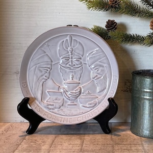 May include: A white ceramic plate with a raised design of three wise men with gifts for the Christ child. The plate is on a black stand and has the text "1975 Gifts for the Christ Child".