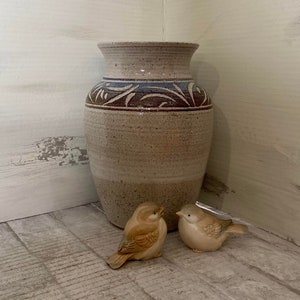 May include: A beige ceramic vase with a decorative blue and brown band near the top. Two small, tan ceramic bird figurines are in front of the vase. The background is a corner with white and gray painted wood.